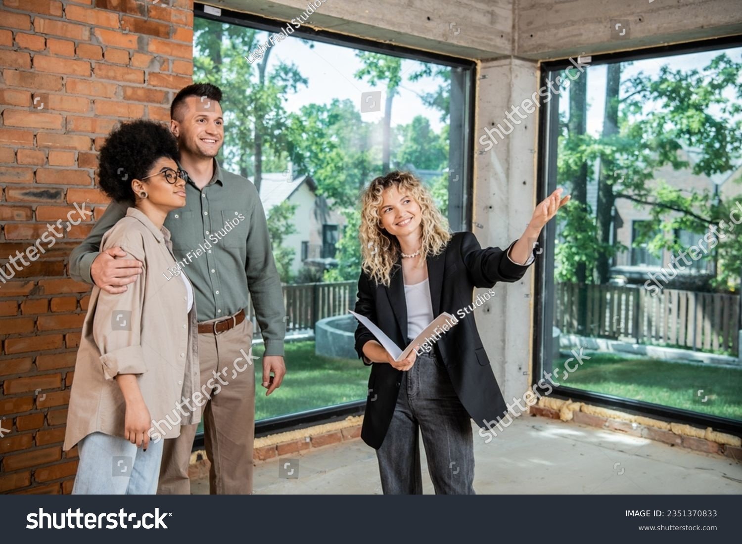 stock-photo-smiling-real-estate-broker-pointing-with-hand-while-showing-new-dwelling-to-multiethnic-couple-2351370833